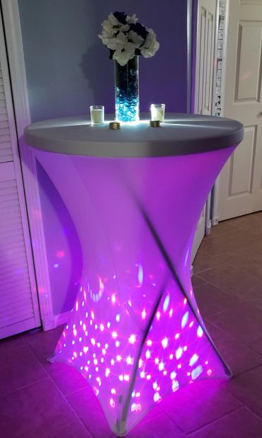 Tall illuminated purple table with flowers and candles on top.