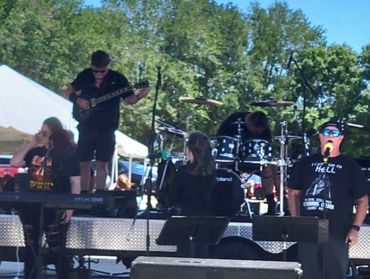 Outdoor band performing live with keyboard, drums, and guitar under clear sky.