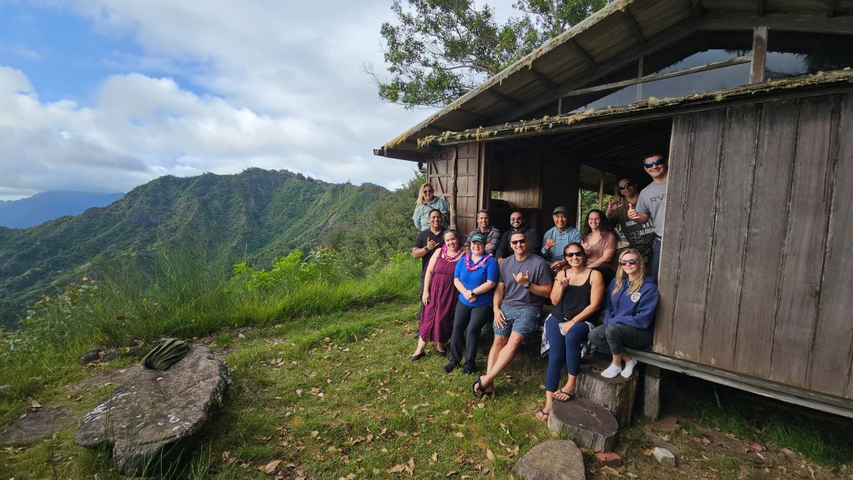 Group of people posing outdoors near a wooden cabin with lush mountains in the background.