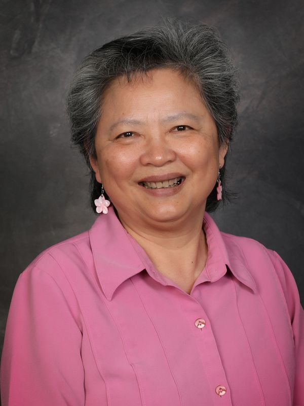 Smiling older woman in a pink shirt with flower earrings against a dark background.