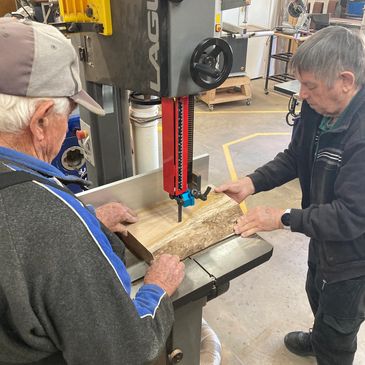 Two elderly men working with a bandsaw on a piece of wood.