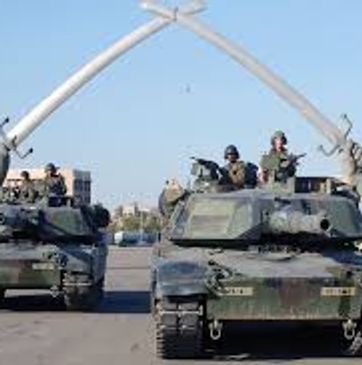 Military tanks with soldiers passing under a large arch structure.