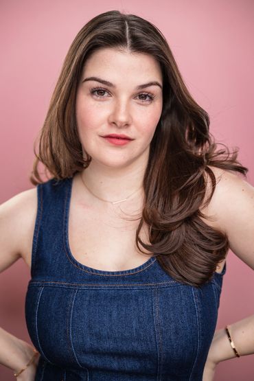 Young woman with long brown hair wearing a denim dress against a pink background.