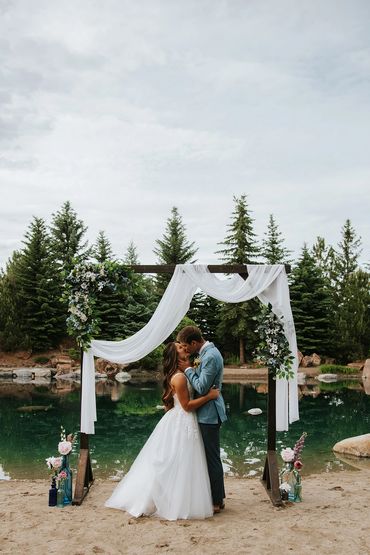 Bride and groom kiss under floral arch by a serene lake.
