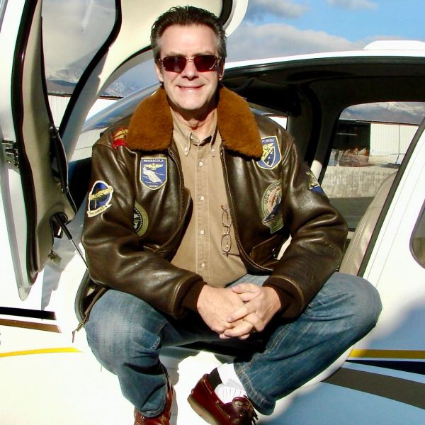 Man in aviator jacket sitting on airplane doorway.