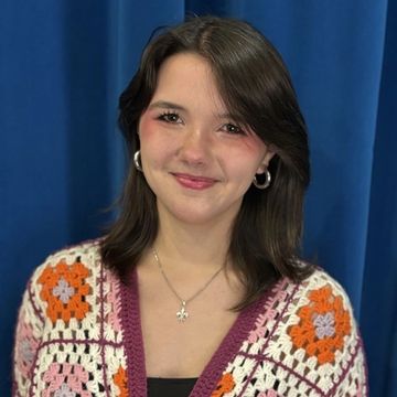 Young woman smiling against a blue backdrop wearing a crocheted cardigan.