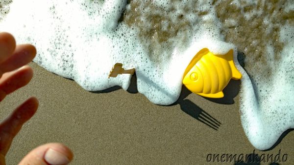 A yellow toy fish on sand with sea foam and a hand reaching out.