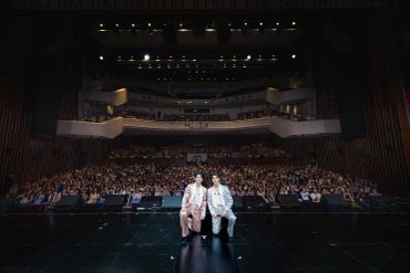 Two performers sit on stage facing a large audience in a theater.