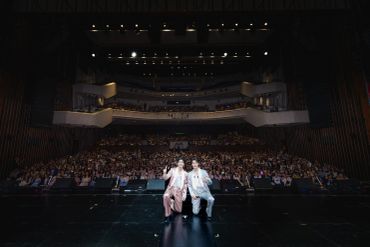 Two performers pose on stage in front of a large, seated audience inside a theater.