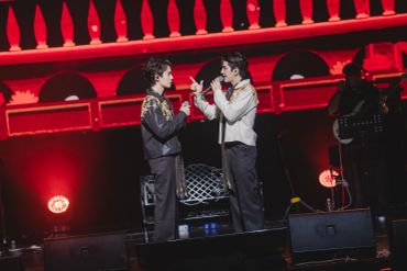 Two performers engaged in a dramatic scene on stage with red lighting.