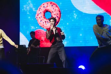 A performer sings energetically on stage with colorful donut visuals in the background.