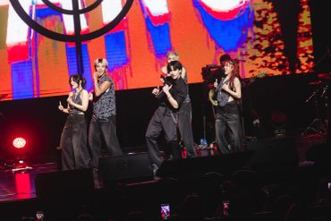 A five-member group performs energetically on stage with vibrant red and blue lights.
