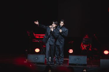 Two performers in black suits singing together on stage with red lighting.