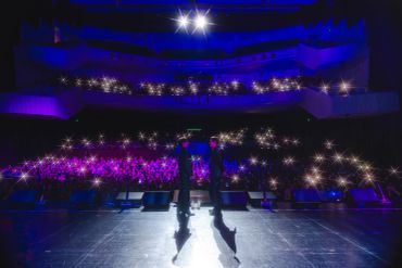 Two performers on stage with audience lights glowing in dark theater.