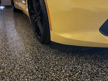 Close-up of a car's front wheel and yellow bumper on a speckled garage floor.