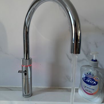 Modern chrome faucet with running water and hand wash bottle beside it.