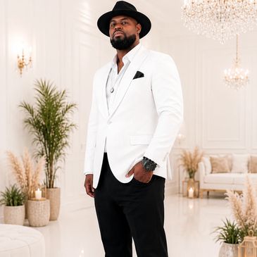 A stylish man in a white blazer and black hat poses confidently indoors.