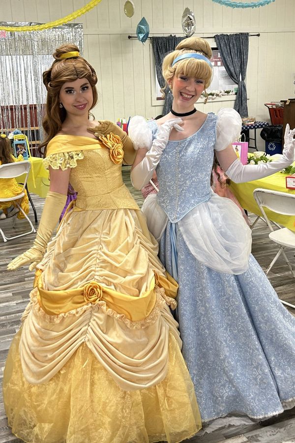 Two women dressed as Disney princesses Belle and Cinderella pose happily indoors.