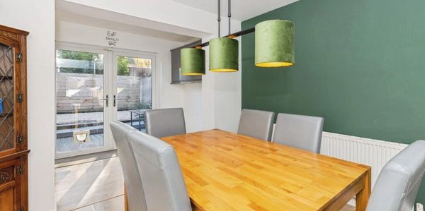 Modern dining room with wooden table, gray chairs, green accent wall, and glass doors leading outside.