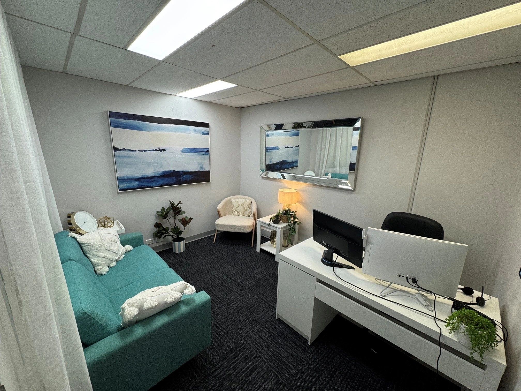 Modern office corner with teal couch, white chair, desk, and wall art.