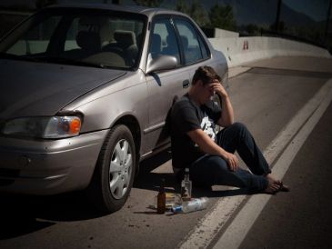 A man sits distressed beside a damaged car with empty alcohol bottles nearby.