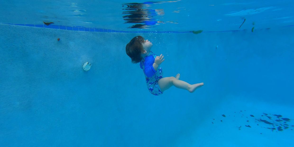 Toddler swimming underwater in a pool wearing a blue swimsuit.