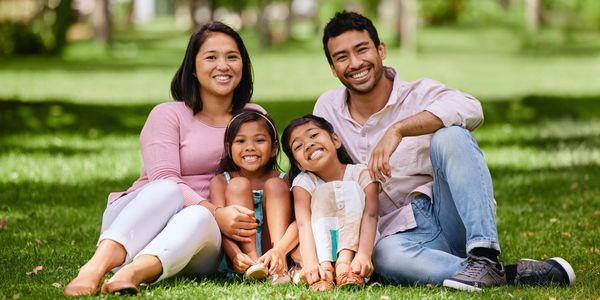 Happy family sitting on grass in a sunny park, smiling at the camera.