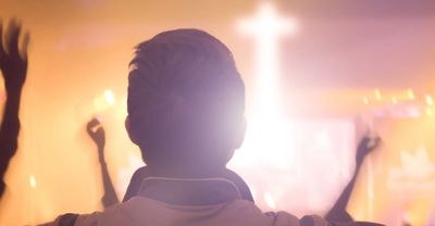 Person with raised hand in front of a glowing cross at a religious event.