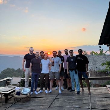 Group of nine men posing on a wooden deck at sunset with mountains in the background.