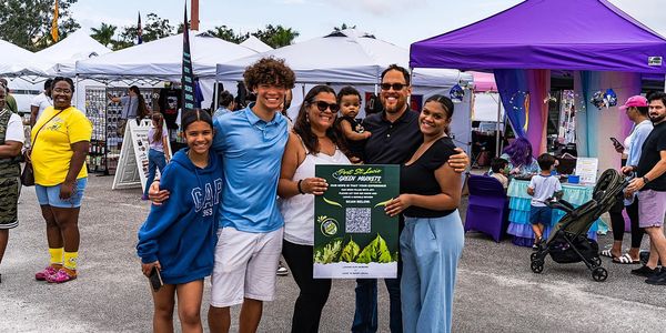 A happy family posing with a Port St. Lucie Green Markets sign at an outdoor event.