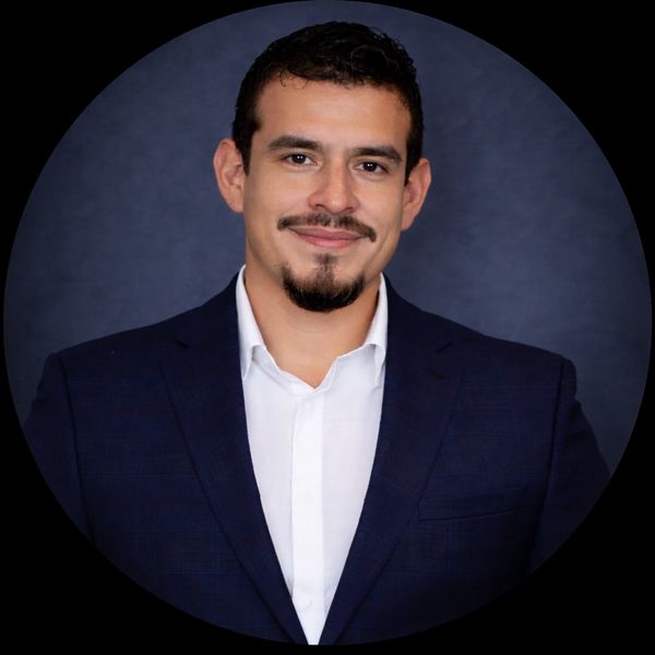 Man in navy blue suit and white shirt smiling confidently against dark background.