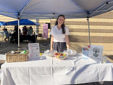 Young woman stands behind a table at an outdoor event booth with snacks and informational signs.