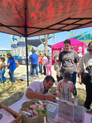 Man helps young girl with an activity at an outdoor event booth under a red canopy.
