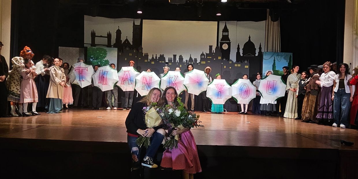 Cast holding umbrellas spelling 'THANK YOU' on stage with two women holding flowers in front.