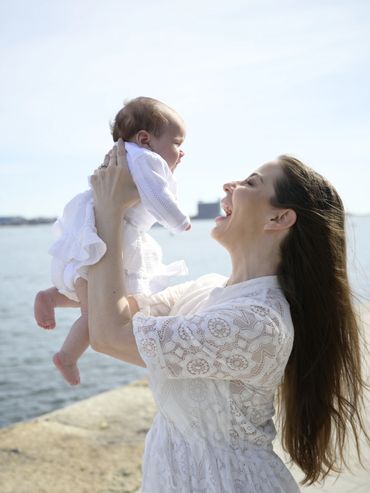 A joyful mother lifts her baby by the water on a sunny day.