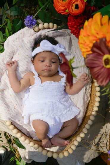 Newborn baby in a white dress lying in a basket surrounded by colorful flowers.