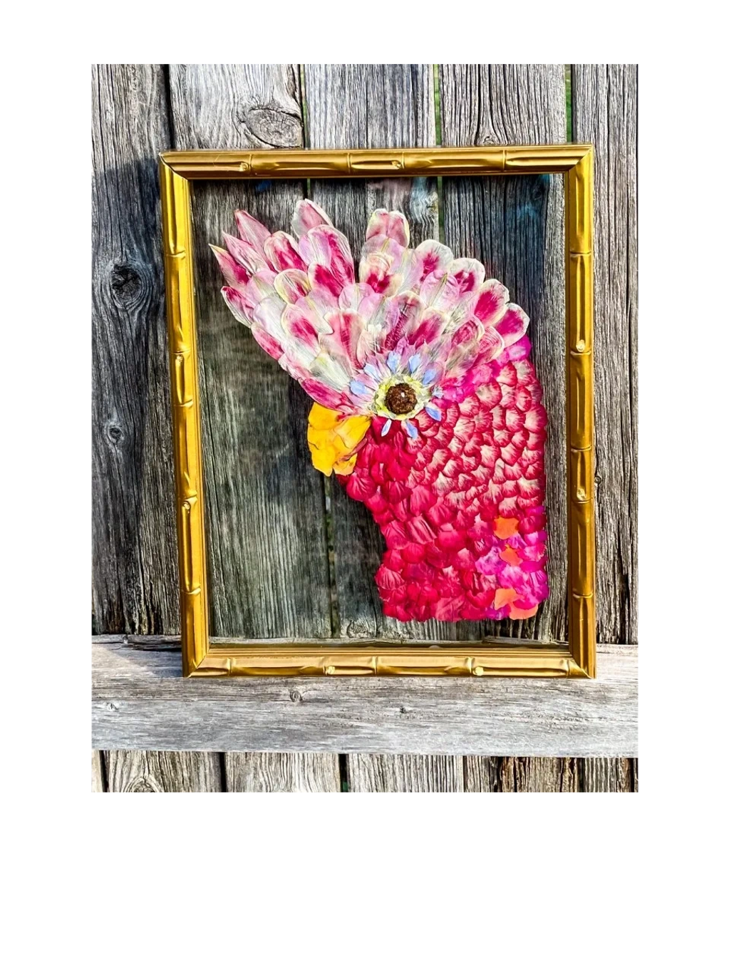 A pink, pressed flower cockatoo, between to pieces of glass in a gold, vintage frame.