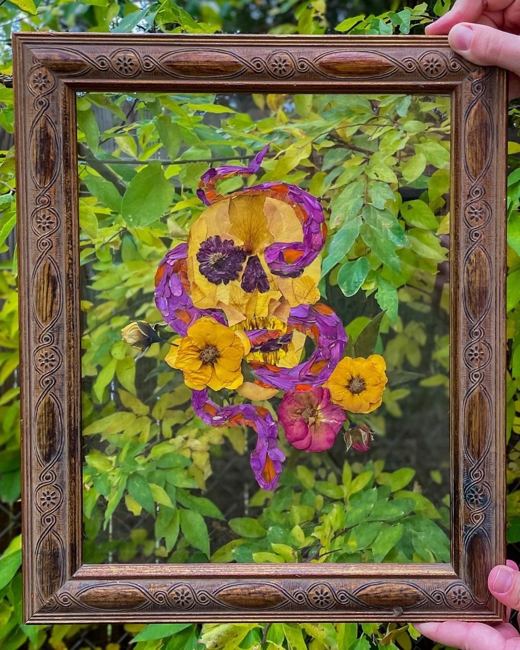A skull and snake made out of pressed flowers, between two pieces of glass in a vintage frame.