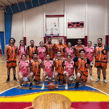 Two basketball teams posing together on court, wearing orange and pink uniforms.