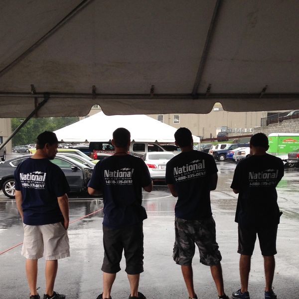 Four men wearing matching National Hail & Dent Removal shirts stand under a tent on a rainy day.