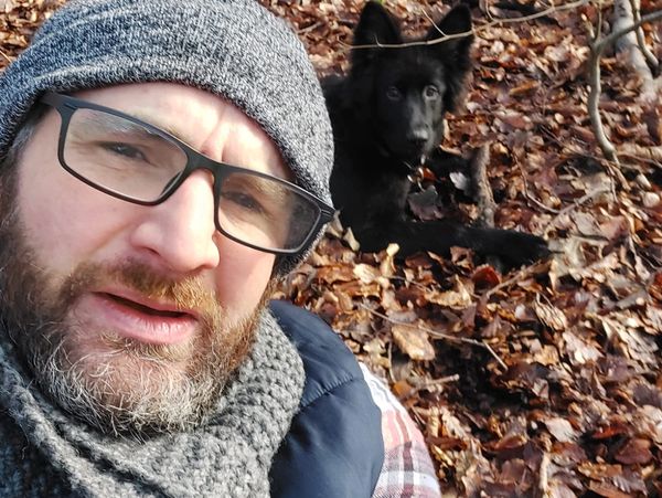 Adam sitting surrounded by autumn leaves with a black German Shepherd in the background