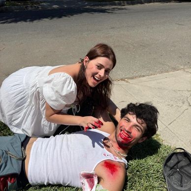 Two people with fake blood, smiling on the ground outdoors.