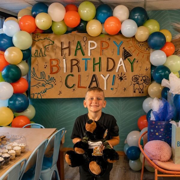 A boy celebrating his birthday with balloons and a teddy bear.