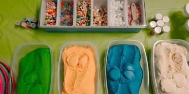 Four containers of colorful kinetic sand with various crafting materials above them.