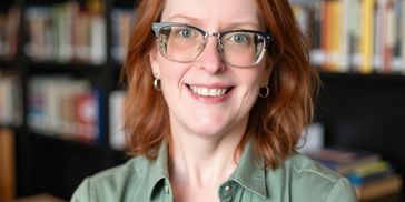 Smiling woman with glasses and red hair in a cozy library setting.