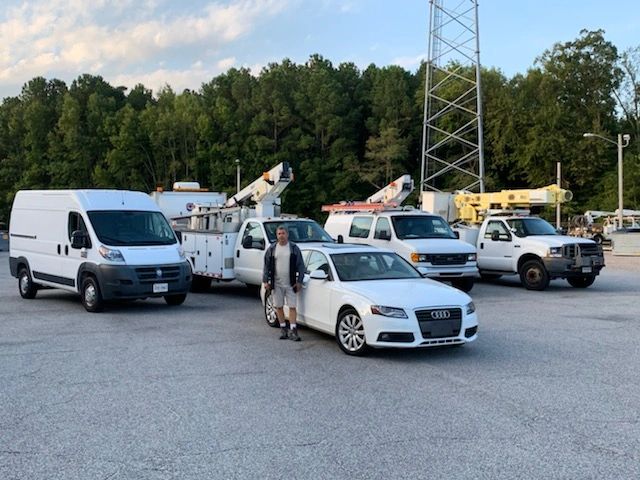Man standing beside a white Audi in front of utility trucks and a van.