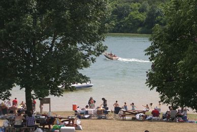 Sandy beach area with people at a picnic and boats in the back.