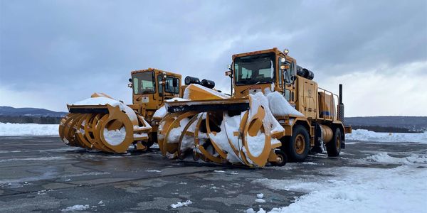 Two large orange snow plows on an RDG runway they recently plowed. Plows are covered in snow.