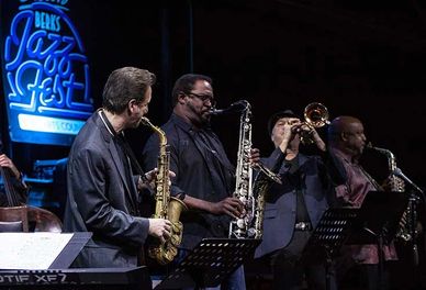 Men in black lined up playing trumpets and saxophones. 