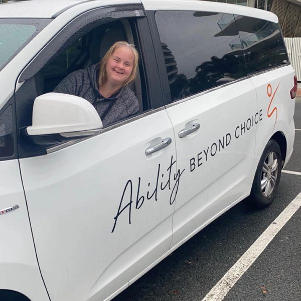 Smiling woman with Down syndrome driving a white van labeled 'Ability Beyond Choice'.